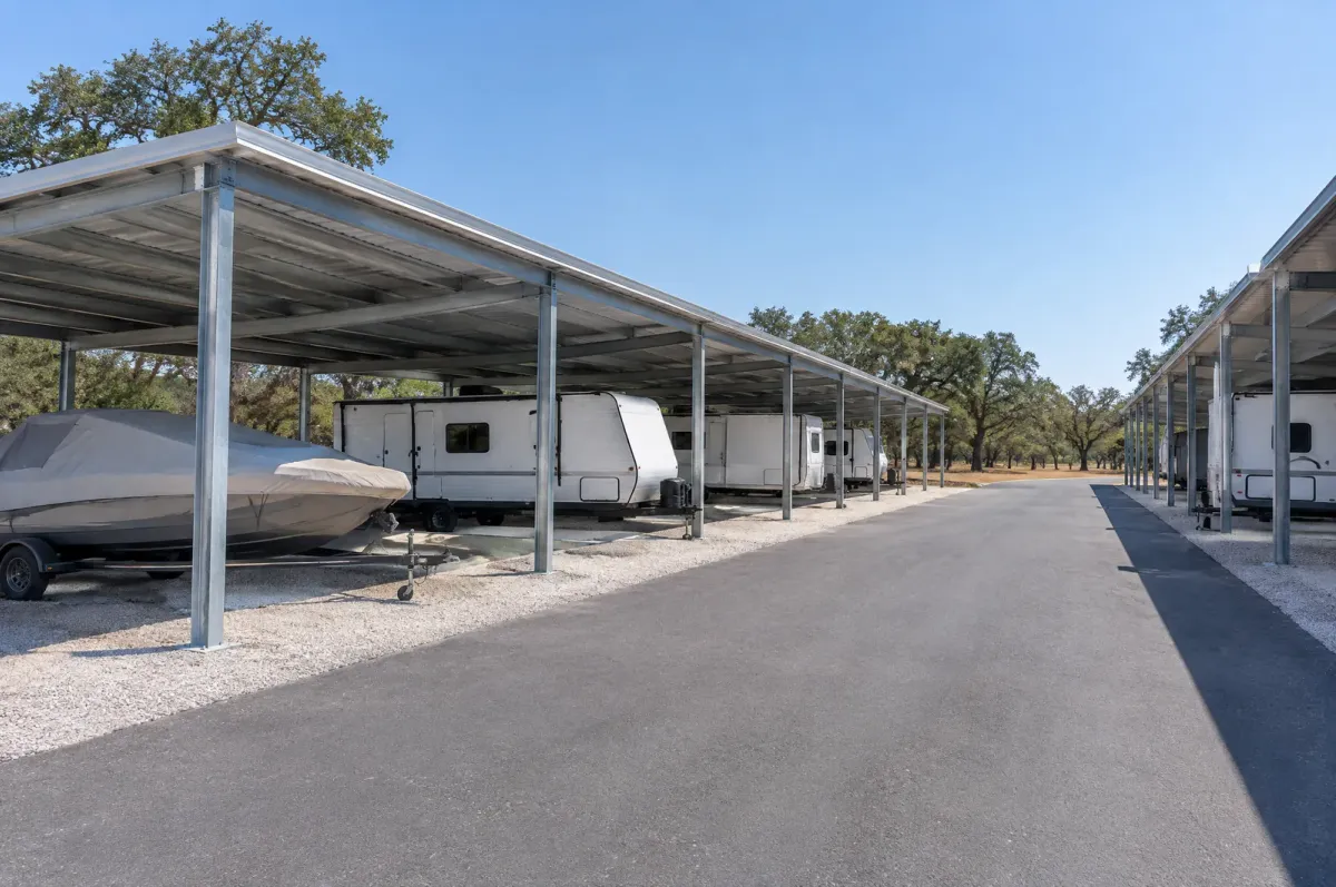 Rows of open steel canopies over stored travel trailers and a covered boat along a paved drive under live oaks